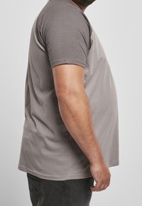 Man som bär en grå t-shirt med kort ärm och mörka byxor, sedd från sidan med höger arm avslappnad längs kroppen.