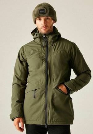 Mężczyzna w zielonej zimowej kurtce z kapturem i wełnianej czapce, z jedną ręką w kieszeni, na jasnym, jednolitym tle.