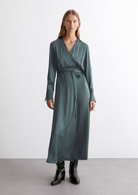 Lang, omslagstil kjole i dyp teal sateng med V-hals, tettsittende ermer og midjebånd. Kombinert med svarte høye støvler.