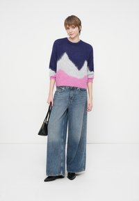 Young person in a blue, white, and pink color-block sweater, wide-leg jeans, holding a black handbag, standing on a white background.