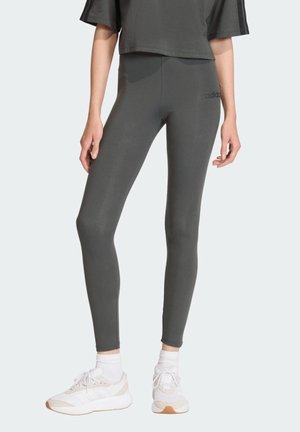 Personne portant des leggings Adidas gris foncé, associés à des chaussettes blanches et des baskets blanches, sur un fond clair.