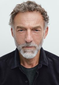 Gråhåret mand med fuld skæg, iført en mørk knap-op skjorte over en mørk T-shirt, har dybtliggende blå øjne og et alvorligt udtryk.