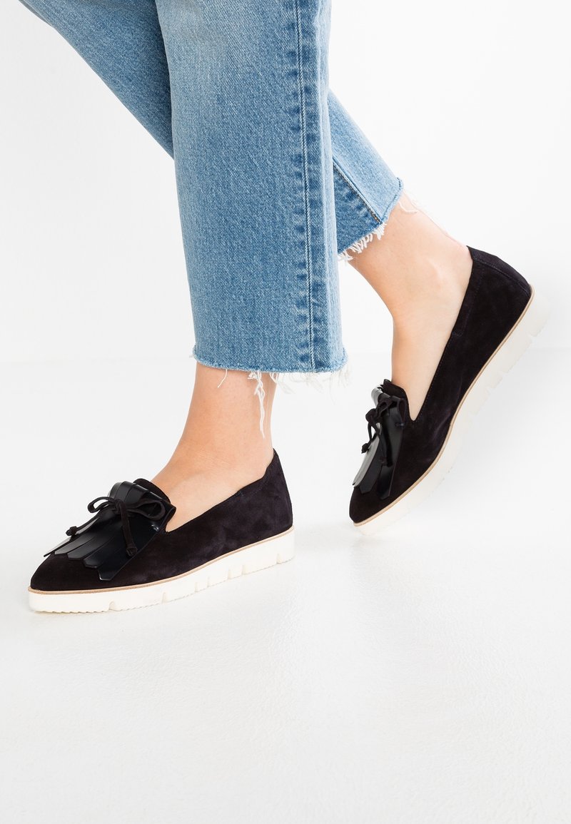 Personne portant un jean court bleu clair et des mocassins en daim noir avec des semelles blanches et un détail à pampilles noires sur un sol blanc.