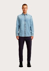 Jasnoniebieska koszula dżinsowa z kołnierzykiem na guziki, długimi rękawami i widocznym przeszyciem, w zestawieniu z ciemnymi spodniami i czarnymi butami.