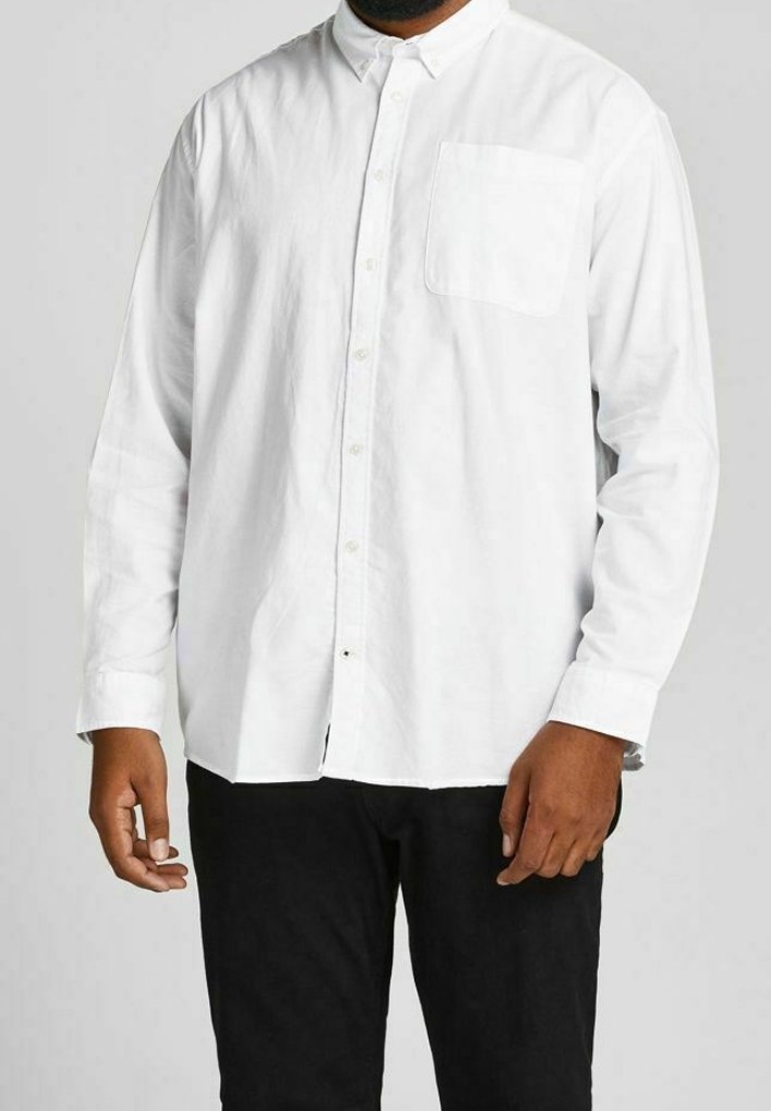 Homme portant une chemise blanche à manches longues boutonnée avec une poche poitrine et un pantalon noir, debout devant un fond uni.