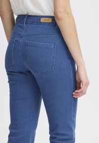Modre jeans hlače z oprtim silhouetom, zadnjimi žepi in oznako na pasu. Tkanina je gladka z rahlim raztezanjem.