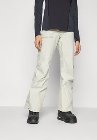 Pantalons imperméables légers de couleur crème avec une coupe droite, dotés d'une poche zippée et d'un ourlet renforcé. Portés avec des bottes noires.
