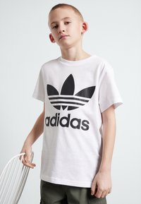 Balts kokvilnas T-krekls ar lielu melnu Adidas logo, kas satur trīs svītras un zieda formas dizainu krūšgalā. Īsas piedurknes un apaļa kakla izgriezums.