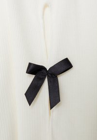 Tessuto a coste crema con un fiocco in satin nero in un'apertura piccola, che evidenzia il contrasto di texture e colore.