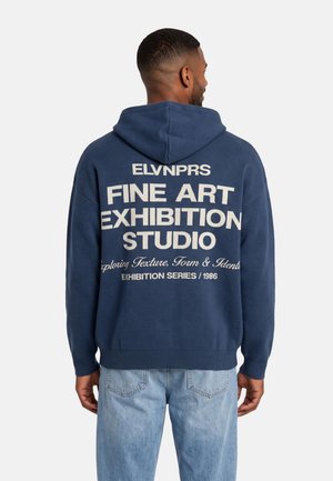 Un hombre visto de espaldas luce una sudadera azul marino con capucha y grandes letras en color crema que dicen «Fine Art Exhibition Studio», combinada con unos vaqueros azul claro.