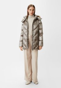 Kurtka puchowa w metalicznej srebrze z kapturem i sztucznym futrem, z ukośnym pikowaniem i zapięciem na zamek, zestawiona z kremowymi spodniami.