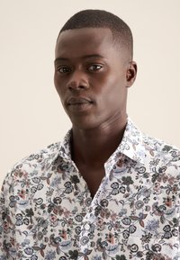 Witte bloemenshirt met een mix van kleurrijke bloemen en bladeren in blauw, roze en groen, met een klassieke kraag en knoopsluiting.