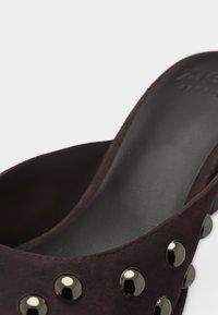 Κοντινή λήψη ενός mule παπουτσιού από σκούρο καφέ σουέτ με μεταλλικούς στρογγυλούς τρουκς κατά μήκος της πάνω άκρης και μαύρη επένδυση με μαξιλάρι.