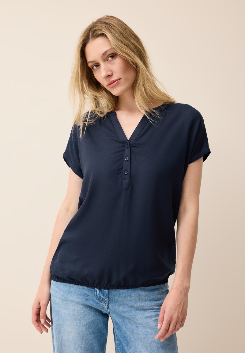 Top blu navy a maniche corte con scollatura a V e bottoni, caratterizzato da una trama liscia e un orlo elasticizzato, abbinato a jeans blu chiaro.