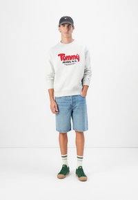 Gray sweatshirt with red "Tommy Jeans" logo, denim shorts, green sneakers with gum soles, and striped socks. Model stands against a neutral backdrop.