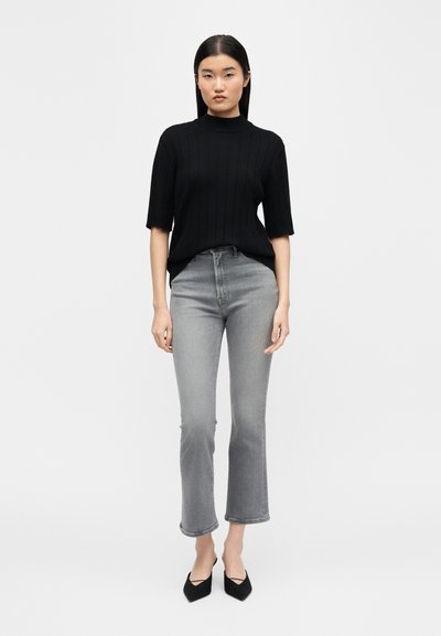 Γυναίκα με ίσια μαύρα μαλλιά που φοράει μαύρο κοντομάνικο ριπ πουλόβερ με ζιβάγκο, γκρι cropped τζιν και μαύρες μυτερές γόβες.