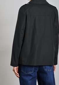 Persona che indossa una giacca nera con spalline a bottone e jeans blu, vista da dietro contro un muro chiaro e semplice.