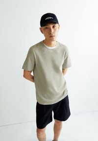 Kurzärmliges, hellgraues T-Shirt mit Rundhalsausschnitt und strukturiertem Stoff, kombiniert mit schwarzen Shorts. Baseballmütze mit Logo, marineblauer Farbe.