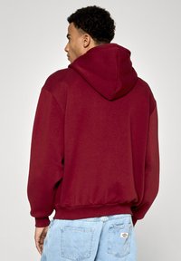 Bordo kapuce ar priekšējo kangoru kabatu, regulējamu auklu kapuce un ribotām aprocēm, apvienota ar gaiši ziliem nolietotiem džinsiem.