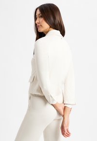 Frau mit langen braunen Haaren, die ein weißes maßgeschneidertes Blazer und passende Hosen trägt, in Seitensicht mit den Händen hinter dem Rücken gefaltet.