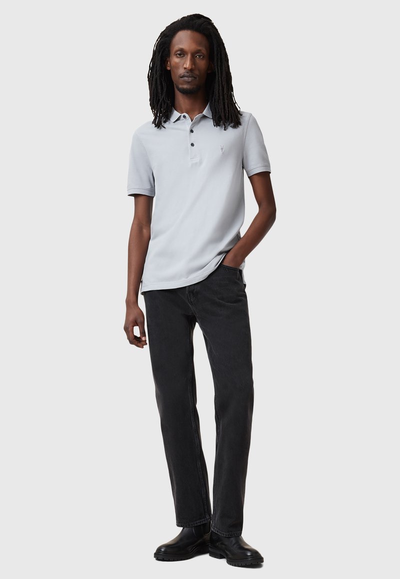 Jasnoszara koszulka polo z krótkim rękawem, z dwu-guzikowym zapięciem, w połączeniu z czarnymi dżinsami i czarnymi butami. Czysty design, minimalne akcesoria.