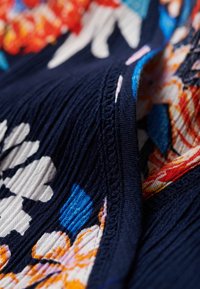 Tessuto blu navy con pieghe testurizzate, caratterizzato da motivi floreali colorati in arancione, blu e bianco. Cuciture visibili che evidenziano i dettagli.