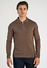 Brun långärmad polo med halv dragkedja och spetsig krage. Har ribbat ärmslut och nederkant, tillverkad av ett mjukt stickat material.