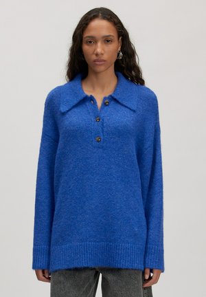 Vrouw met lang donker haar, gekleed in een felblauwe gebreide trui met drie knopen en een brede kraag, gecombineerd met versleten zwarte jeans.