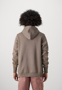 Sweat à capuche marron avec capuche à cordon, poignets côtelés et ceinture. Le modèle le porte avec un pantalon cargo rose clair.