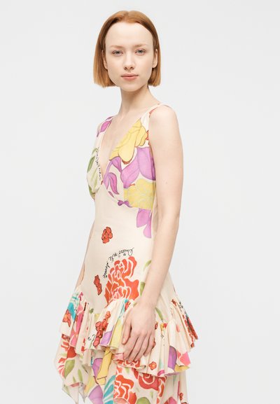 Νέα γυναίκα με κοντά κόκκινα μαλλιά που φοράει αμάνικο φόρεμα με ανθισμένο σχέδιο και βολάν στο τελείωμα, πάνω σε απλό λευκό φόντο.