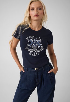 Mujer rubia con camiseta negra de Guess con un gran logo y vaqueros de talle alto en azul oscuro, de pie con las manos en los bolsillos.