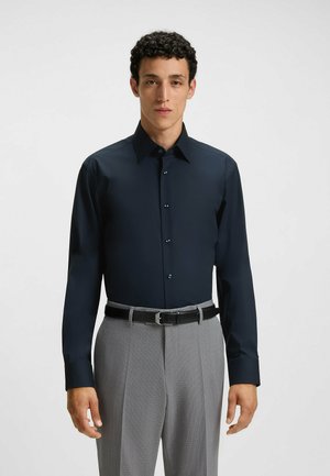 Marineblaue Hemd mit spitzem Kragen und Knopfleiste. Verfügt über dunkle Knöpfe und lange Ärmel. Getragen mit grauen gemusterten Hosen und schwarzem Gürtel.