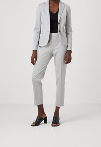 Kobieta w jasnoszarej, dopasowanej marynarce i dopasowanych, skróconych spodniach, czarnym topie bez rękawów oraz czarnych, otwartych na palce sandałach na obcasie, stojąca.