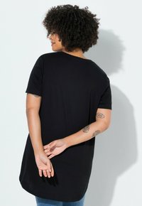 Femme aux cheveux bouclés portant un t-shirt noir à manches courtes et un jean bleu, debout les mains jointes derrière le dos, de profil.
