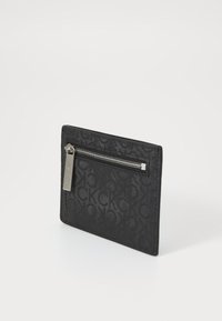 Portefeuille noir avec poche zippée, présentant une surface texturée avec un motif en monogramme, des finitions en métal et une forme rectangulaire fine.