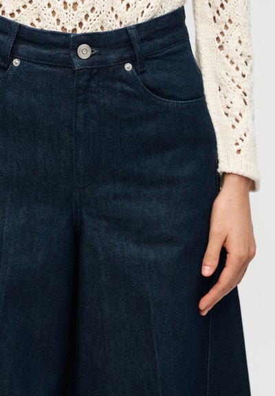 Κοντινό πλάνο σκούρων μπλε τζιν παντελονιών στερεωμένων με μεταλλικό κουμπί, συνδυασμένα με κρεμ πλεκτό πουλόβερ και ένα εμφανές χέρι στο πλάι.