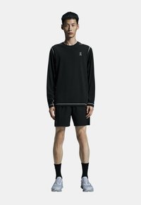 Sort atletisk langærmet skjorte med hvid syning og logo, parret med sorte shorts. Båret med sorte sokker og grå løbesko.