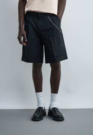 Hombre que lleva pantalones cortos negros a medida hasta la rodilla con cadena lateral, calcetines blancos y mocasines negros sobre un fondo liso.