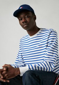 Blauw-wit gestreepte blouse met lange mouwen, voorzien van een rode borduring van "Parijs", gecombineerd met een marineblauwe pet met een Franse vlag-embleem.