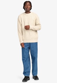 Beige kabelgebreide trui met een ronde halslijn, gecombineerd met losse blauwe denim jeans en zwarte schoenen. De trui heeft ribbels bij de mouwen en de onderrand.