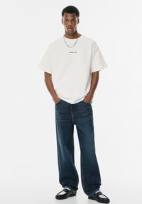 Afslappet beige t-shirt med "ERROR CODE" tekst, kombineret med mørkeblå vide bukser og sorte sko. Enkel, afslappet påklædning.