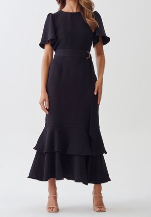 Czarna sukienka z krótkimi falbaniastymi rękawami, dopasowaną talią i falbanistym, warstwowym dołem. Materiał wydaje się gładki, z paskiem ozdobionym okrągłą sprzączką.