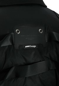 Chaqueta negra con una textura suave y mate; cuenta con detalles de ojetes y una etiqueta con la inscripción "MWM Design" cosida en la parte de atrás.