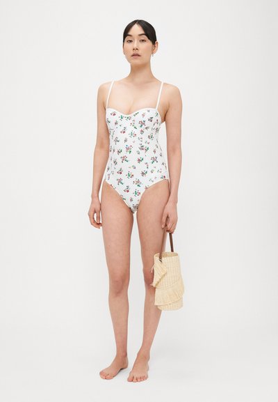 Costume intero bianco a fiori con spalline sottili. Presenta uno scollo a cuore e un design aderente. La modella tiene una borsa di paglia con frange.