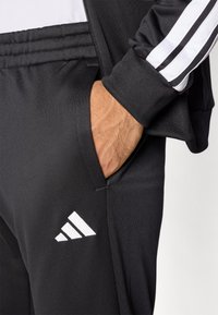 Čierne športové nohavice s bočným vreckom, na ktorých je biele logo Adidas. Tkanina je hladká s zúženým strihom. Detaily tvoria biele pruhy.