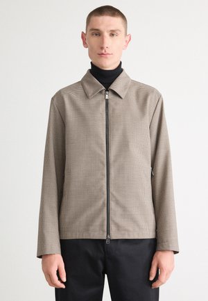 Chaqueta de cuadros marrón con cuello y cremallera frontal. Mangas largas y textura suave, utilizada sobre un cuello de tortuga negro.