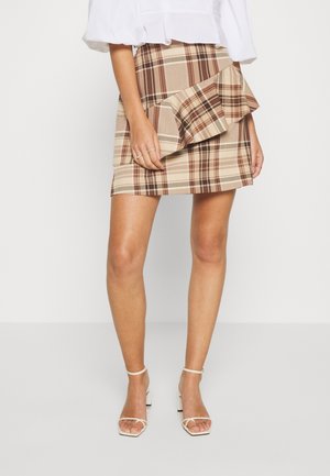Femme portant une mini-jupe à carreaux beige et marron avec un détail volanté, un chemisier blanc et des sandales blanches à talons avec brides.