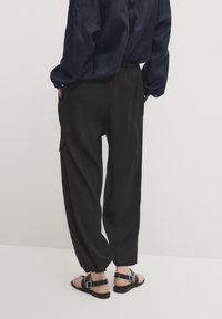 Musta, väljät housut, joissa on kapenevat lahkeet, joustava vyötärönauha ja sivutaskut, yhdistetty mustiin remmikengännauhoihin. Kankaan pinta on teksturoitu.