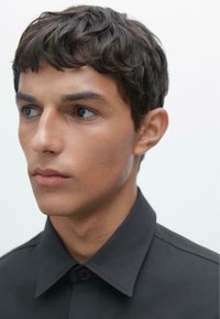 Black shirt with a button-up collar, featuring a smooth fabric texture. The garment is worn by a person with short, dark, wavy hair.