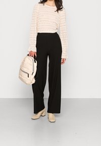 Haut à manches longues beige avec motif en dentelle, pantalon noir à jambes larges, chaussures à talons carrés beiges et un sac à dos beige avec détail de fermeture éclair.